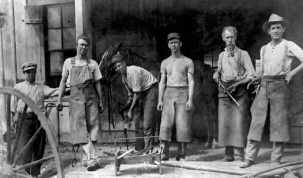 Historical photo of six blacksmiths outside a workshop, showcasing tools and attire from the past.