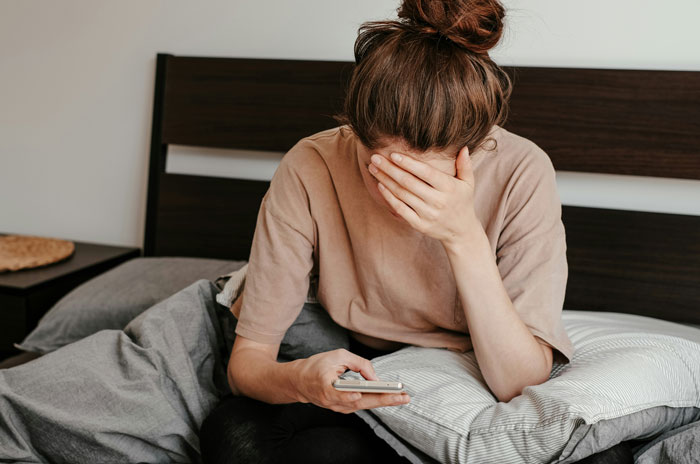 Woman sitting on a bed with a phone, appearing distressed, highlighting mental health struggles.