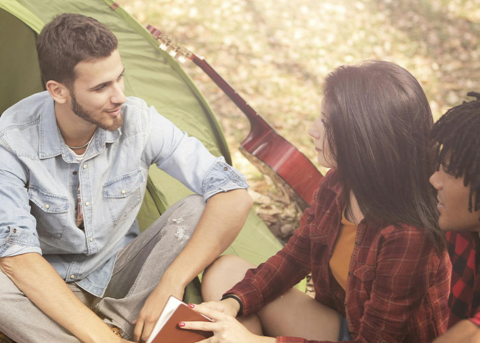 Three friends sit by a tent, sharing supportive reactions about coming out, with a guitar in the background.
