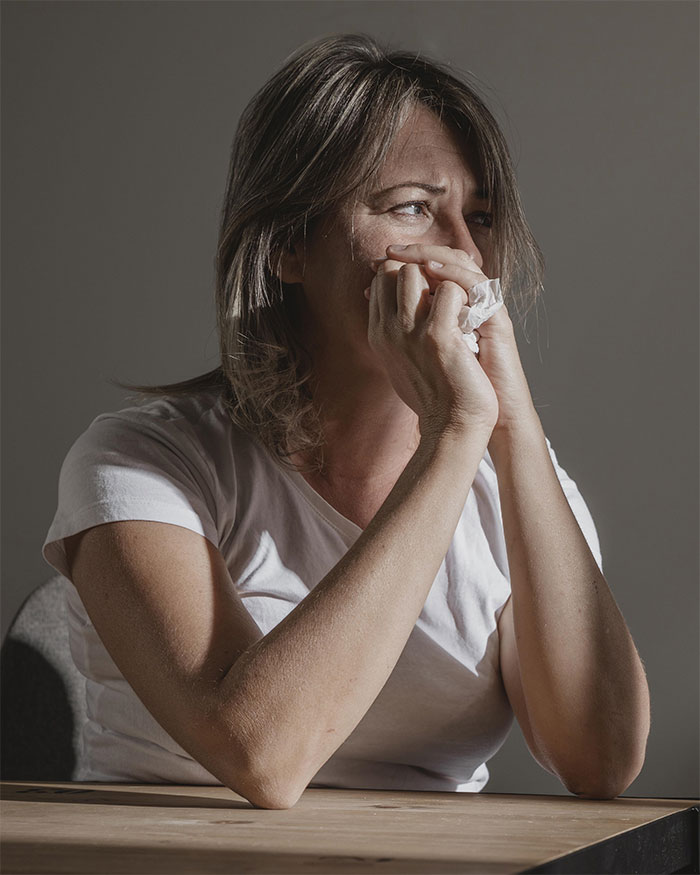 A woman in a white shirt looking thoughtful, holding a tissue, with natural light highlighting her expression.