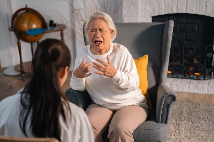 Grandmother in a white sweater gesturing emotionally while seated in a living room setting.