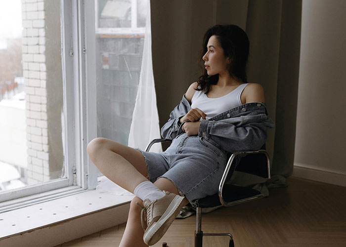 Person in denim outfit sitting on chair by a window, embodying modern beauty tips.