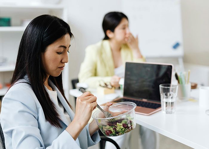 Woman eating salad at desk, looking concerned, with laptop and coworker in background, illustrating a toxic workplace.