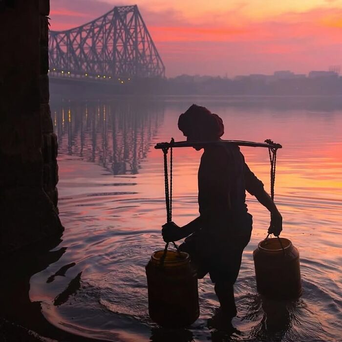 Silhouette of a person carrying water by the river at sunrise with a bridge in the background, capturing an unexpected moment.