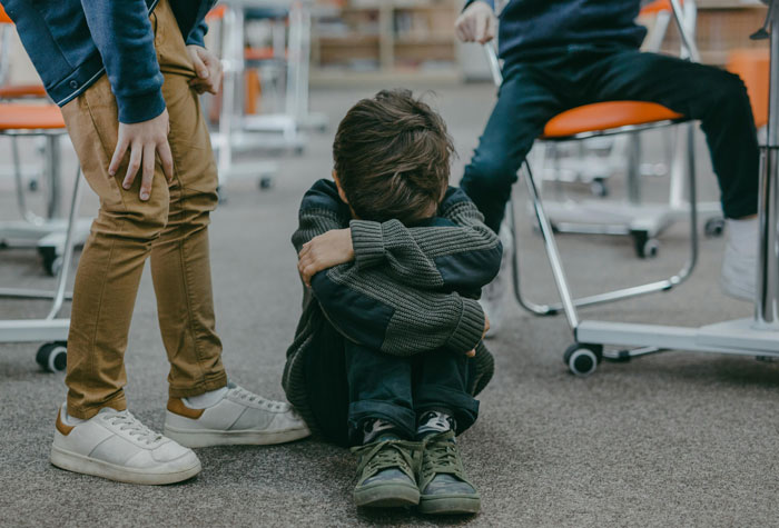 Young person sitting on the floor, hugging knees in distress, with two others nearby in a classroom.