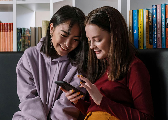 Two friends comfortably seated, happily looking at a phone, sharing reactions to coming out stories.