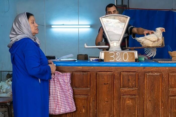 Woman holds a bag, observing a vendor weighing a chicken at a market; an unexpected street photo moment.