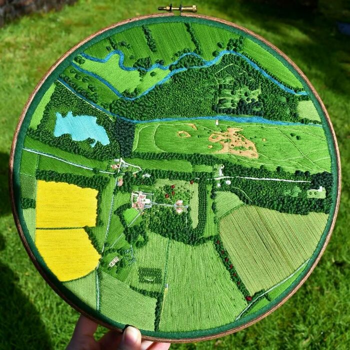 Landscape embroidery art, showcasing intricate green fields and blue rivers, held in a wooden hoop.