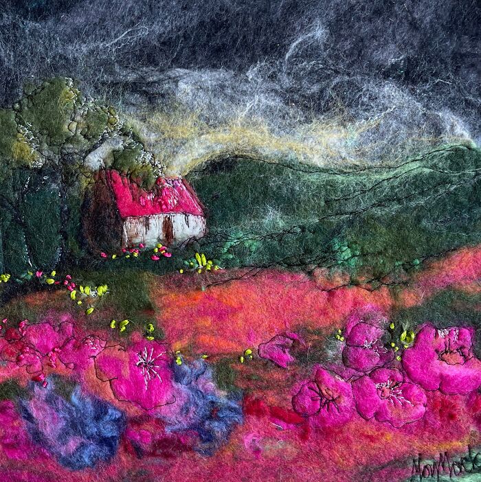 Fiber art depicting a vibrant landscape with a small red-roof cottage amidst pink flowers and green hills.