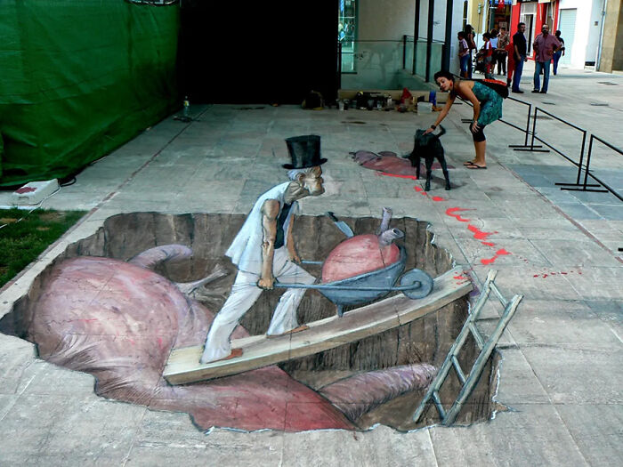 3D street art of a man in a top hat pushing a wheelbarrow on a bridge illusion, with a woman and a dog nearby.