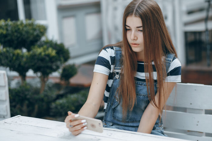 Young woman in denim overalls using smartphone, contemplating normal social habits.