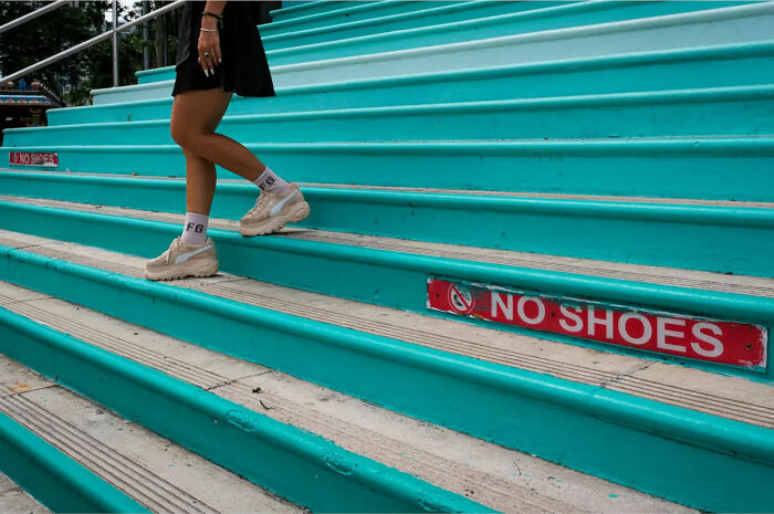 Person in sneakers on turquoise stairs, "No Shoes" sign visible, capturing street life's raw beauty.