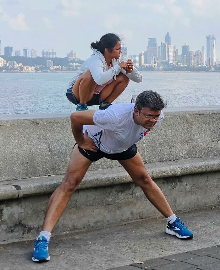 Woman perfectly balanced on a crouching man's back on a street, capturing an unexpected moment with a city skyline in the background.
