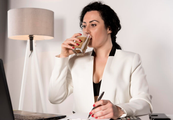 Woman enjoys a drink at her desk, wearing a white blazer, perfect setting for harmless April Fools’ pranks.
