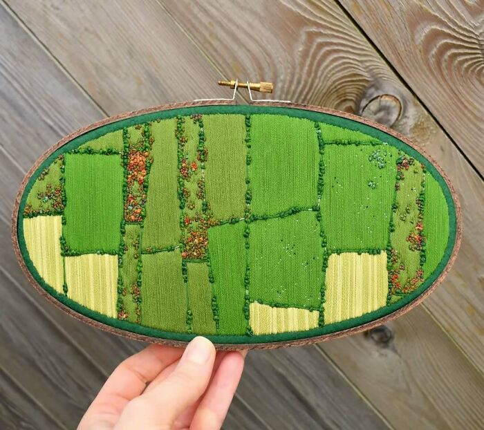 Oval-shaped landscape embroidery depicting fields in green and yellow hues, held by a hand.