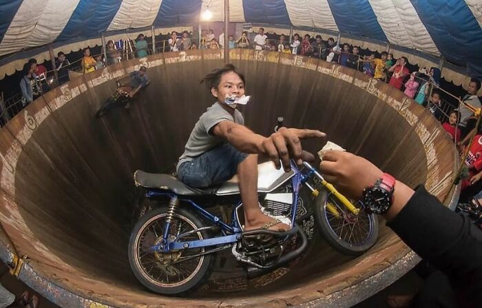 Motorcyclist in a circus ring, performing a perfectly timed trick, surrounded by an audience, capturing an unexpected moment.