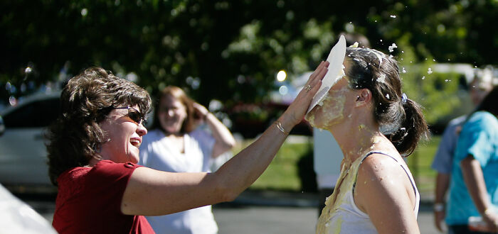 Woman playfully smashes a pie in another woman's face outdoors, surrounded by laughing friends.