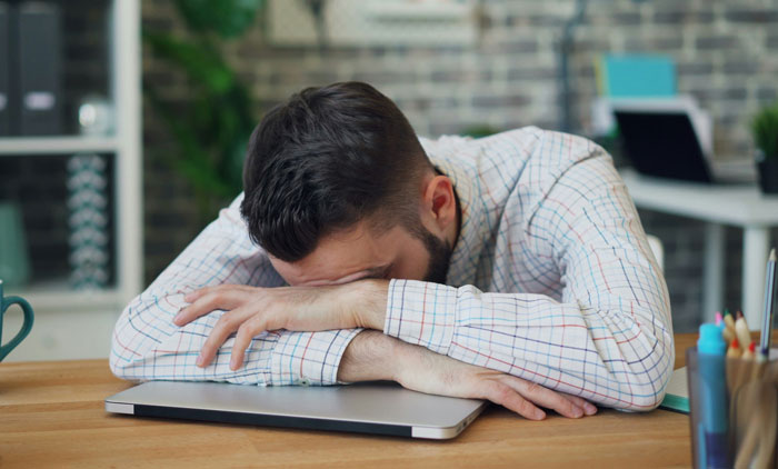 A frustrated man in a plaid shirt rests his head on a laptop, waiting for karma at work.