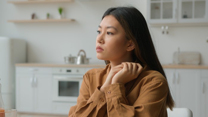 Young woman in a brown shirt sits thoughtfully in a modern kitchen, pondering wedding attire etiquette.