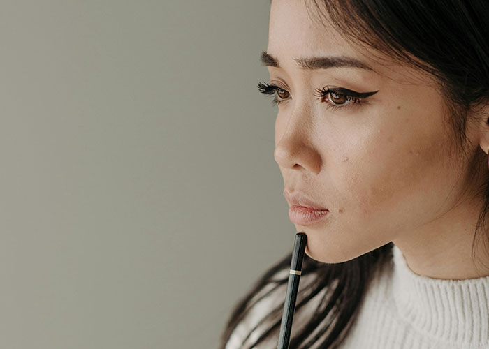 Woman in deep thought holding a pen, wearing a white sweater, contemplating property investment.