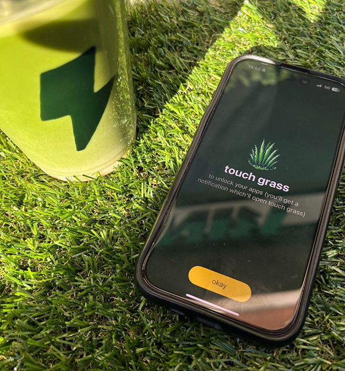 Smartphone on grass displays "touch grass" app, next to a green cup, illustrating social media unblocking method.
