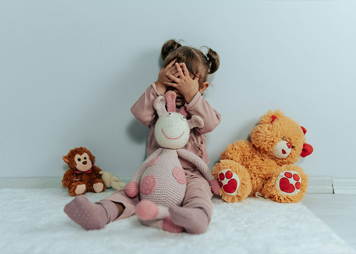 Toddler covering face, sitting with stuffed animals, illustrating behaviors of a trashy parent.