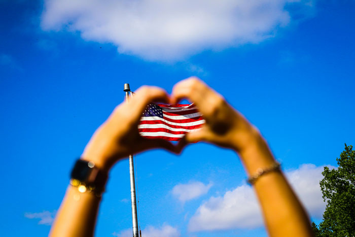 Hands forming a heart shape in front of an American flag under a clear blue sky.