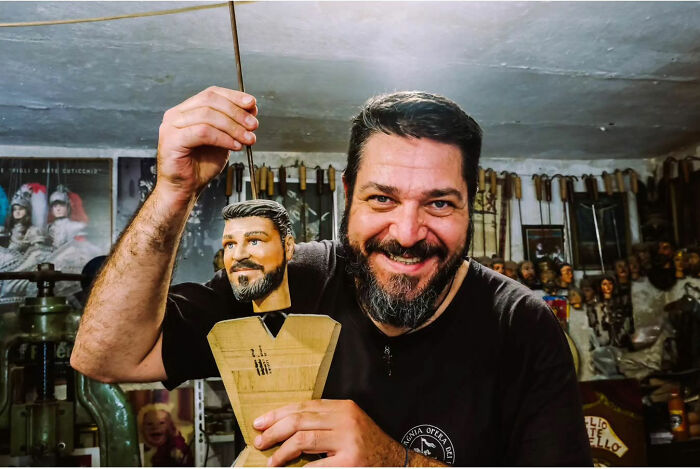 Man smiling while holding a crafted wooden puppet in a workshop, showcasing street life's raw beauty.