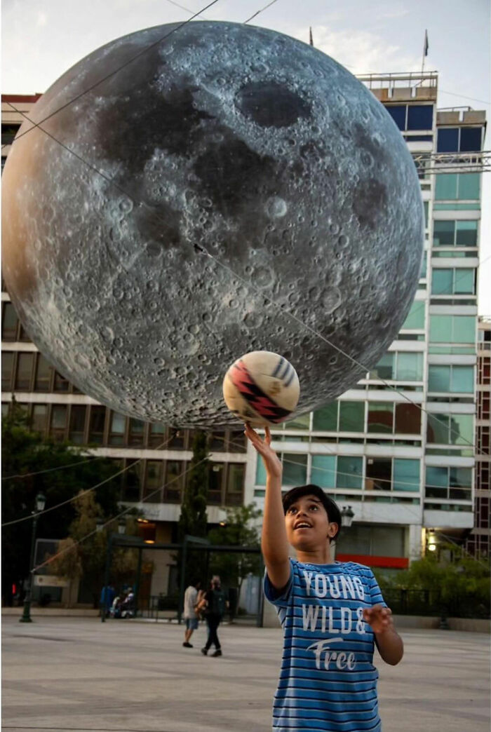Child playing with a ball beneath a large moon sculpture in an urban setting, capturing the raw beauty of street life.