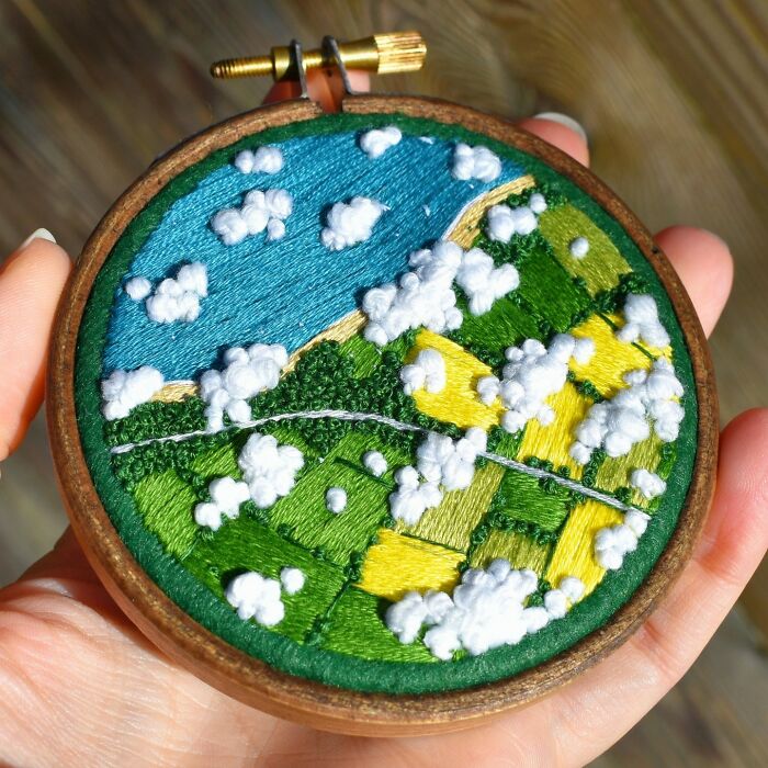 Hand holding a detailed landscape embroidery with clouds, fields, and water.