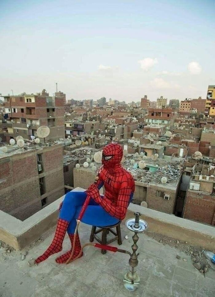 Spider-Man seated on a rooftop, relaxing with a hookah against a cityscape backdrop, embodying random funny moments.