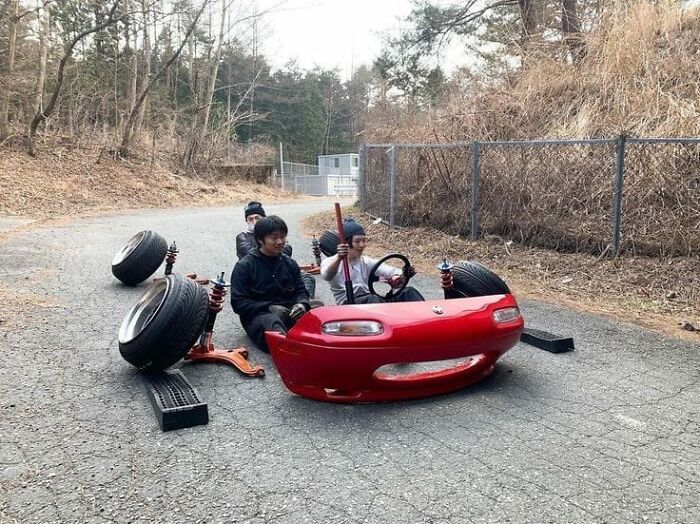 Three people enjoy a humorous ride in a modified, wheel-less car mock-up on a quiet road.