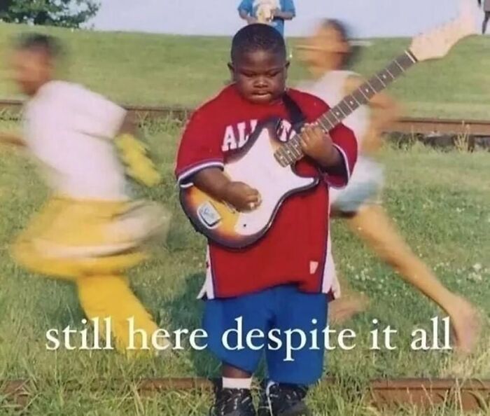 A child holding a guitar with text, "still here despite it all," creating a funny meme moment.