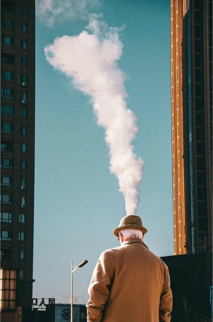 Man in coat and hat stands between buildings with steam above, exemplifying raw street life beauty.