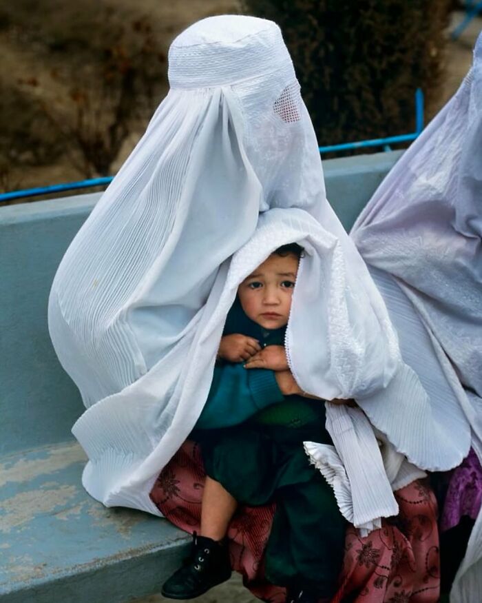 Child peeking out from under a white garment on a bench, capturing an unexpected street moment.