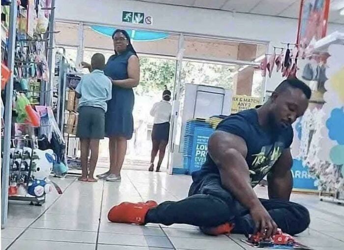 Muscular man in a store aisle, playing with toy cars, while shoppers browse nearby.
