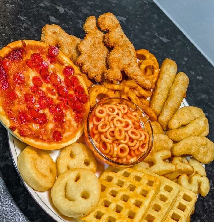 A plate of random funny foods: pizza, smiley fries, dinosaur nuggets, baked beans, and waffles.