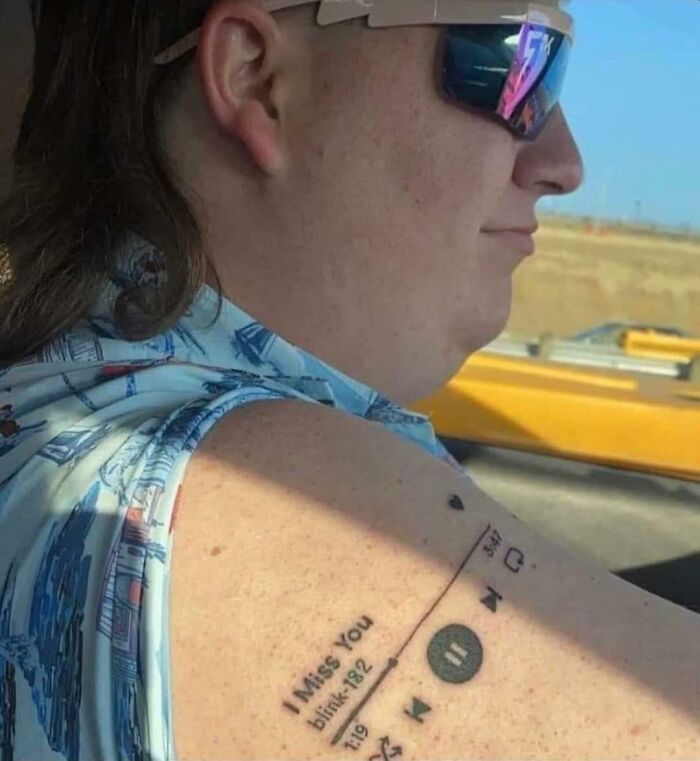 Person with mullet and sunglasses displaying a music player tattoo on their arm, featuring "I Miss You" by Blink-182, showcasing humor.