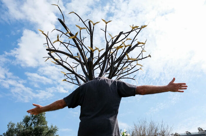 Man with outstretched arms stands before tree sculpture under blue sky, capturing the raw beauty of street life.