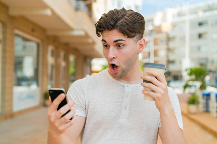 Man in white shirt looks surprised at phone, holding a cup, illustrating what seems normal because we pretend.