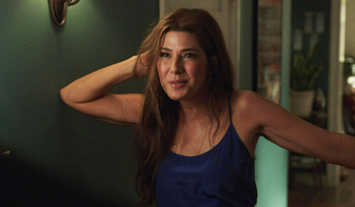 A woman in a blue top smiling and leaning against a wall, engaging in a casual conversation.