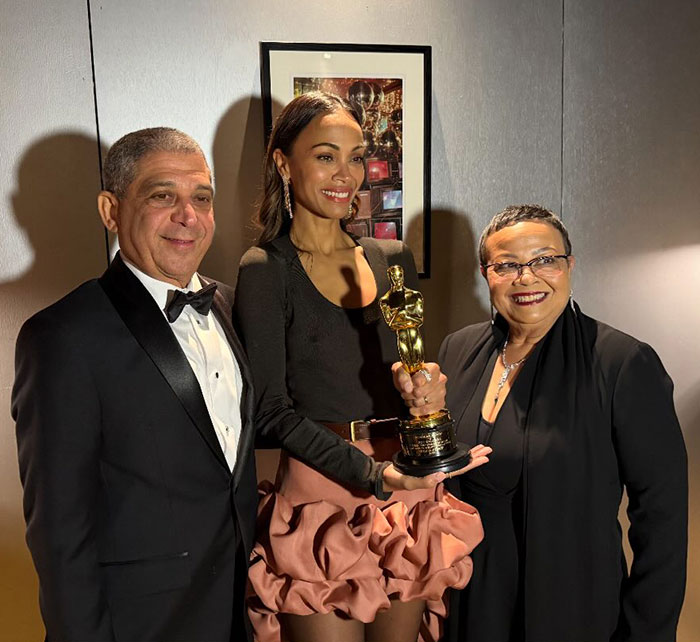 Three people posing with an Oscar award, dressed in formal attire, highlighting a recent event response.