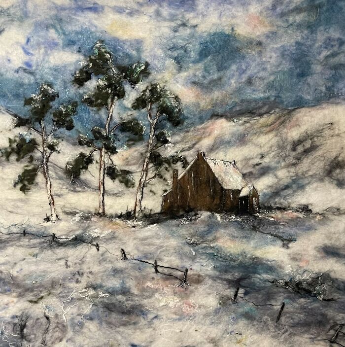 Fiber art landscape by Moy Mackay featuring snowy scenery with trees and a cottage under a cloudy sky.