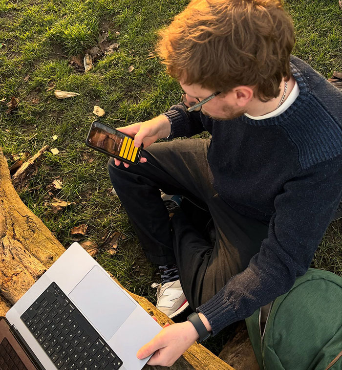 A person using an app on their phone outdoors to unblock social media, sitting on grass beside a laptop.