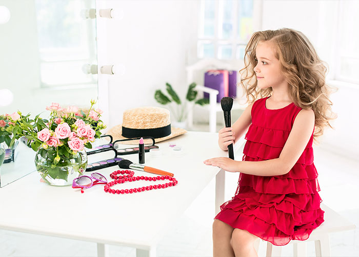 Child with makeup in a bright room, holding a brush, surrounded by accessories and flowers on a vanity table.