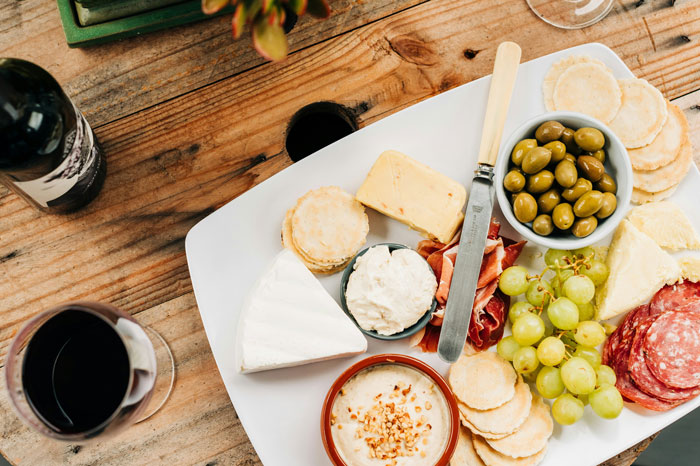 Lazy meal with cheese, olives, grapes, and charcuterie on a platter, accompanied by wine on a wooden table.