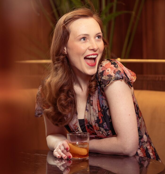 Smiling woman in floral dress holding a drink, embodying wedding joy in an awkward moment.
