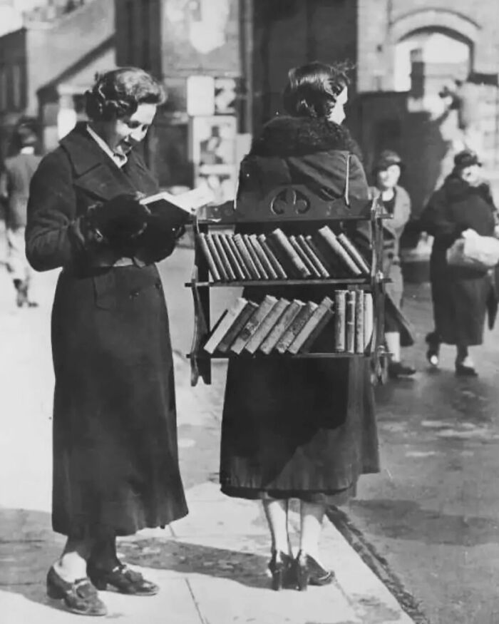 Two women interact with a mobile bookshelf on the street; a perfectly timed street photo capturing an unexpected moment.