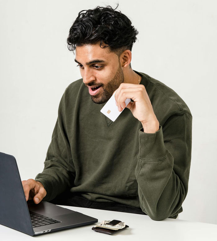Man in green sweater using laptop, holding a credit card, possibly booking a non-refundable vacation. Man in green sweater using laptop, holding a credit card, possibly booking a non-refundable vacation.
