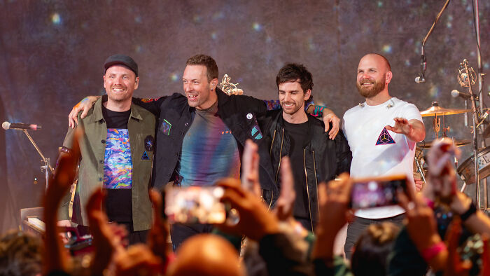 A band on stage, four members smiling and embracing, with fans capturing the moment, exemplifying popularity despite criticism.